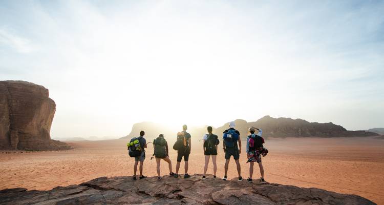 Silhouettenhafte Gruppe von Rucksacktouristen, die auf einem Felsgrat stehen und den Sonnenaufgang über der Wüste Wadi Rum beobachten.