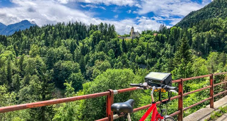 Rotes Tourenrad lehnt an einem Geländer mit Blick auf dichten Kiefernwald und entfernten Kirchturm.