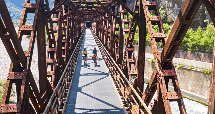 Radfahrer überqueren eine dramatische rostrot-metallic Eisenbahnbrücke hoch über einer felsigen Flussschlucht.