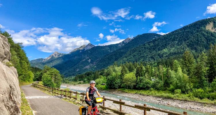 Einsamer Radfahrer auf gepflastertem Weg folgt einem funkelnden Alpenfluss, der von bewaldeten Gipfeln gesäumt wird.