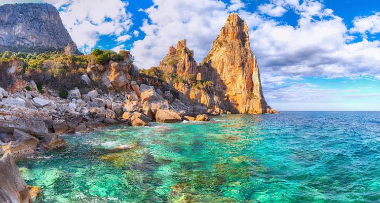Pinnacle calcaire dramatique s'élevant d'une côte accidentée avec une eau turquoise cristalline sous un ciel bleu vif et des nuages épars.