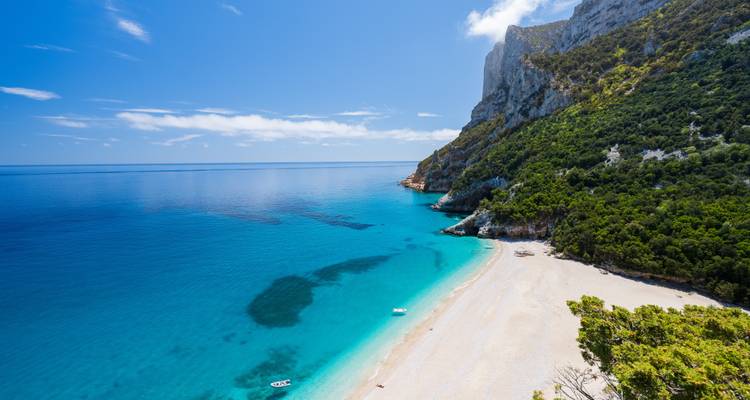 Vue aérienne panoramique d'une plage sardinienne isolée avec du sable blanc, une eau aux dégradés turquoise et des falaises imposantes en arrière-plan.