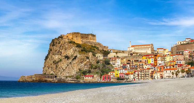 Un pintoresco pueblo costero con una colina rocosa, playa de arena y edificios coloridos.