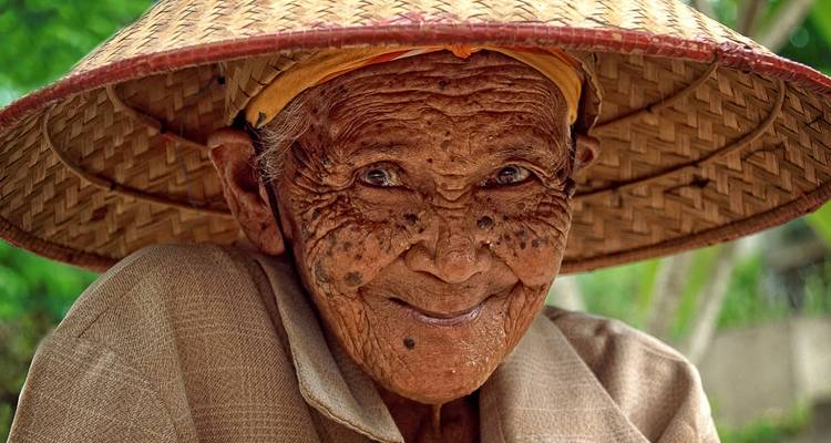 Elderly person smiling and wearing traditional woven hat.