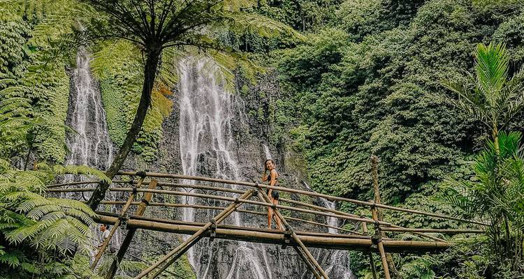 A person on a bamboo bridge with waterfalls surrounded by lush foliage.