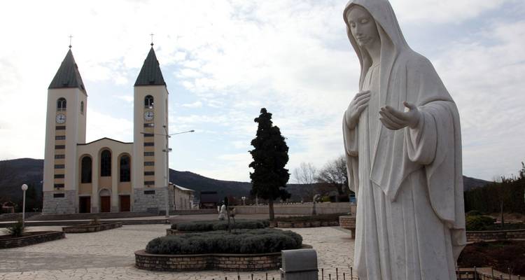 Estatua de mármol blanco de la Virgen María de pie frente a una iglesia de dos torres en una plaza tranquila