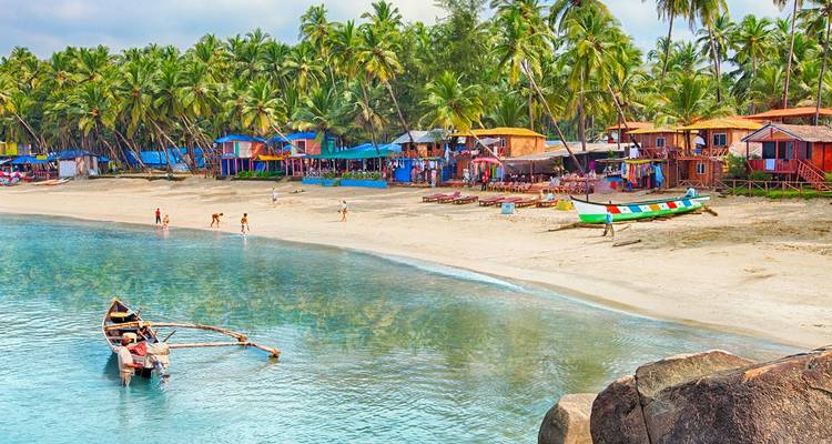 Cabañas coloridas y palmeras bordean una tranquila costa de Goa donde botes de madera flotan en aguas cristalinas.