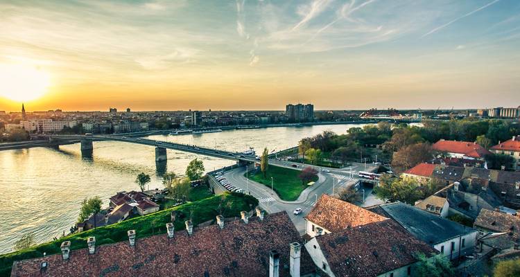 Vista elevada del atardecer sobre una ciudad ribereña con un puente cruzando agua brillante y un cielo dorado y cálido.