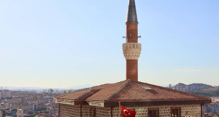 Pequeña mezquita de ladrillo con minarete esbelto que domina un paisaje urbano turco, bandera nacional ondeando en el techo.
