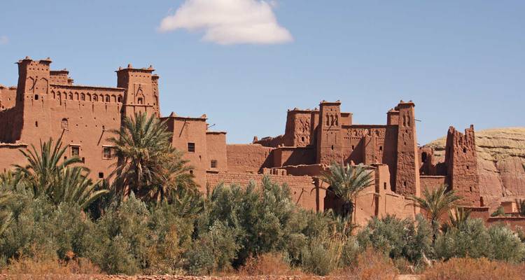 Historic earthen kasbah of Ait Benhaddou rising above palm groves under a blue sky.
