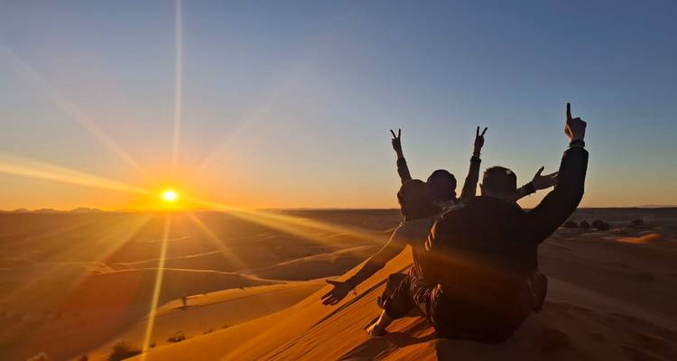 Three friends sit on a dune raising victory signs while watching a vibrant Sahara sunset.