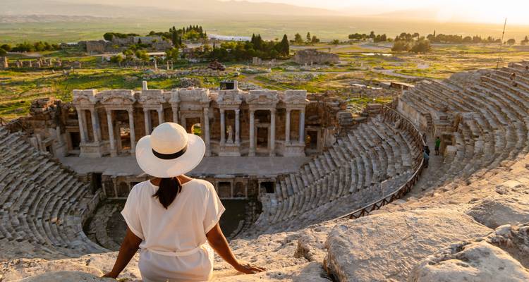 Vrouwelijke reiziger in witte jurk en hoed zittend op steen van Hierapolis theater, uitkijkend over de oude arena tijdens het gouden uur.