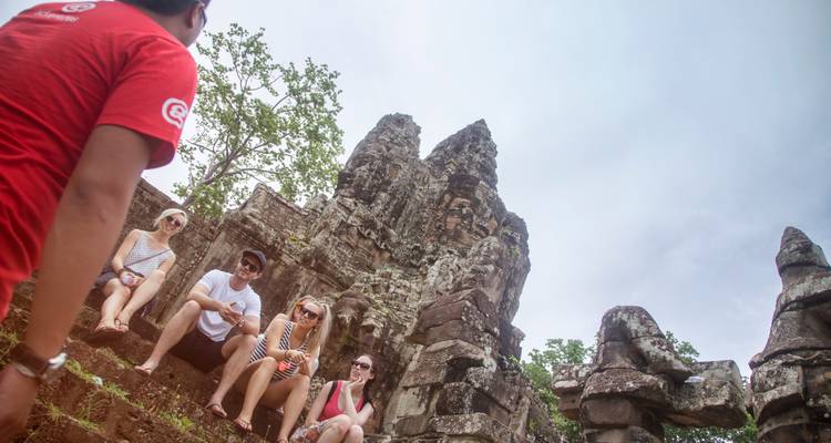 Group of travelers sitting on ancient stone steps listening to guide at Angkor ruins.
