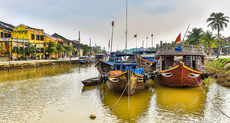 Row of colourful wooden boats tied along Hoi An’s calm river with heritage houses behind.