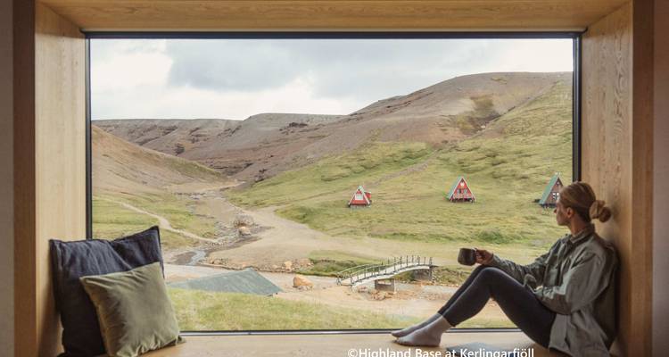 Una persona mirando por una ventana hacia un valle pintoresco con pequeñas cabañas.