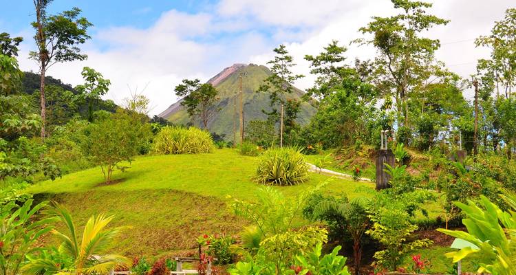 Verdant gardens lead up to a conical volcano peak under a partly cloudy sky.