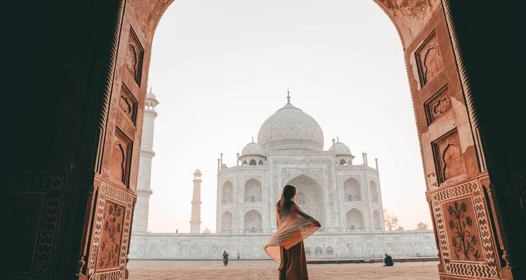 Una mujer envuelta en un chal contempla el Taj Mahal suavemente iluminado desde el arco de una mezquita al amanecer.