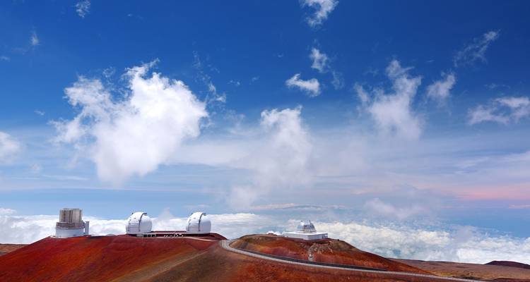 Les dômes blancs des télescopes des Observatoires de Mauna Kea perchés sur le sommet volcanique rouge au-dessus d'une mer de nuages.