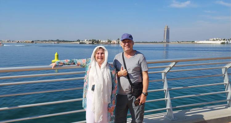 Two people standing on a boardwalk by a body of water, with some distant structures visible.