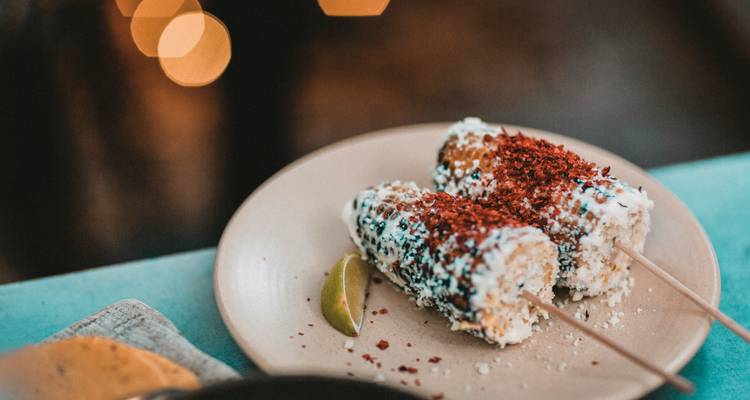 Close-up of two Mexican street corn skewers coated with cheese and chili on a ceramic plate with a lime wedge.