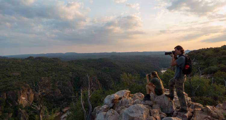 Dos fotógrafos están de pie en el borde de un acantilado rocoso capturando un vasto valle verde y un río sinuoso al atardecer.