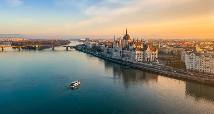 Vue aérienne au coucher du soleil du Parlement hongrois sur le Danube avec un bateau de croisière en dessous.