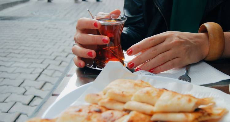 Gros plan d'une personne tenant un verre de thé turc à côté d'une assiette de pâtisseries dans un café en plein air.
