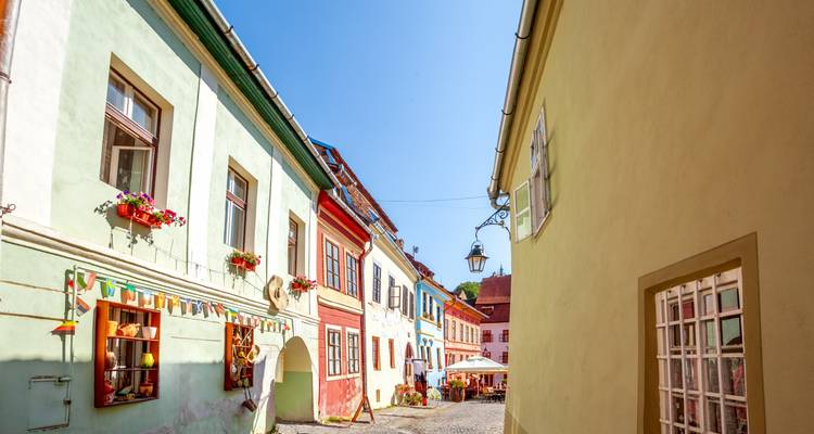 Une rue pavée ensoleillée bordée de maisons aux couleurs pastel et de jardinières fleuries.