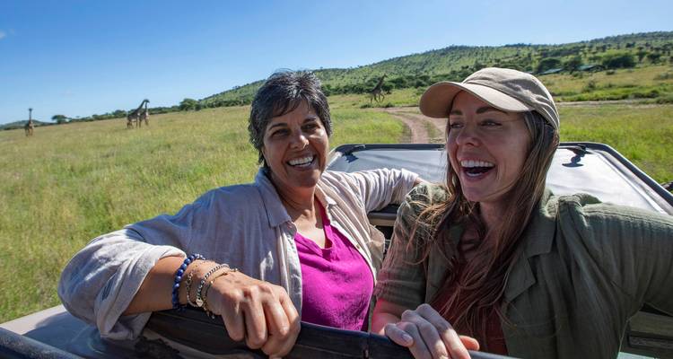 Two smiling women in a safari vehicle with giraffes in the background.