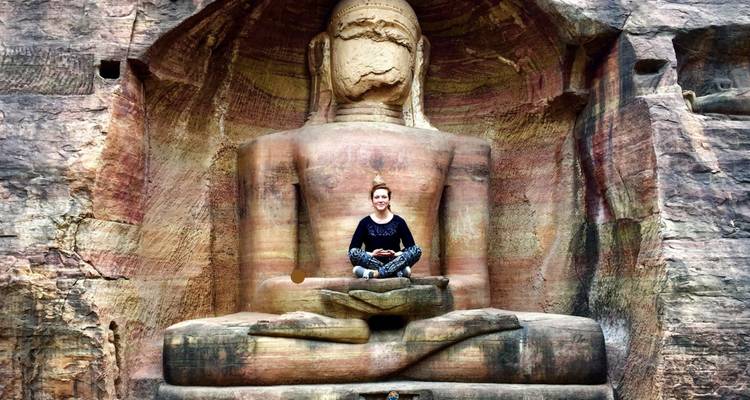 Person sitting in a meditative pose in front of a cliffside carved Buddha statue.