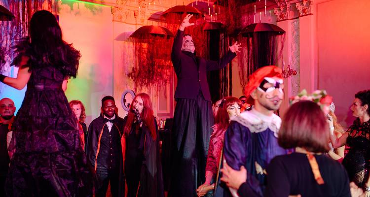 Tall performer on stilts raising arms among masked guests at a lively Halloween celebration.