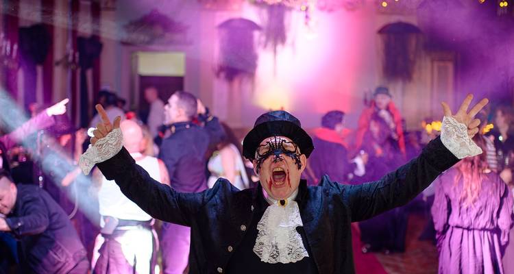 Man in gothic costume shouting with arms wide amid fog and lights at a costumed dance party.