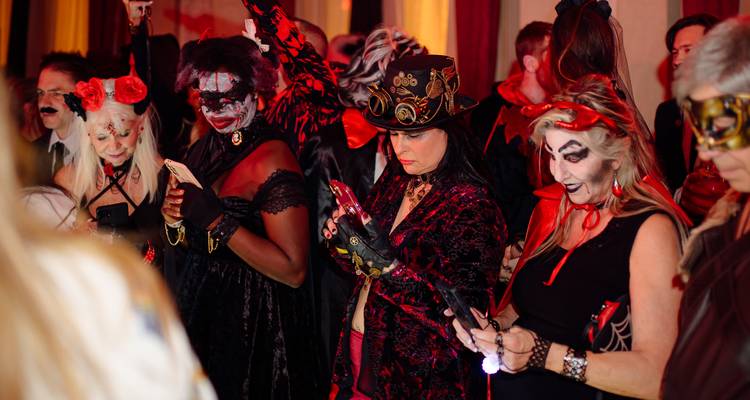 Group of elaborately dressed Halloween attendees checking phones under red lighting.