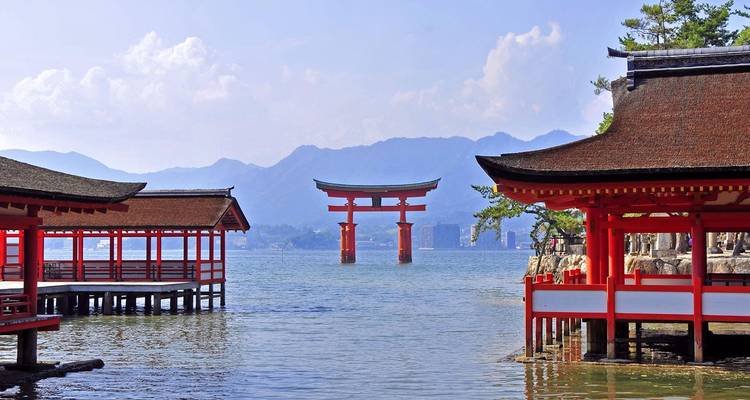 Iconic red torii gate of Itsukushima Shrine rising from the sea framed by shrine pavilions and distant mountains.