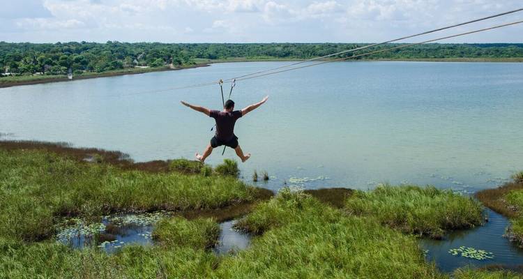 A person zip-lining over a grassy area with a water body below.