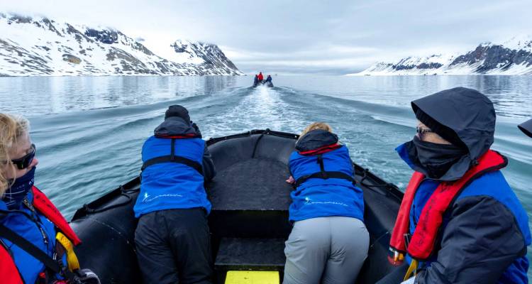 Les passagers vêtus de parkas bleus se penchent sur un zodiac noir qui file à grande vitesse dans un fjord polaire glacé flanqué de montagnes aux sommets enneigés.