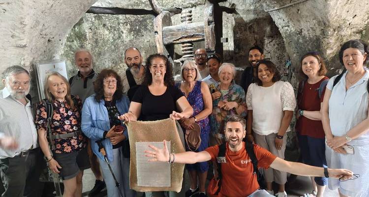 A tour group poses inside a rustic stone olive mill, smiling and holding a sample tray.