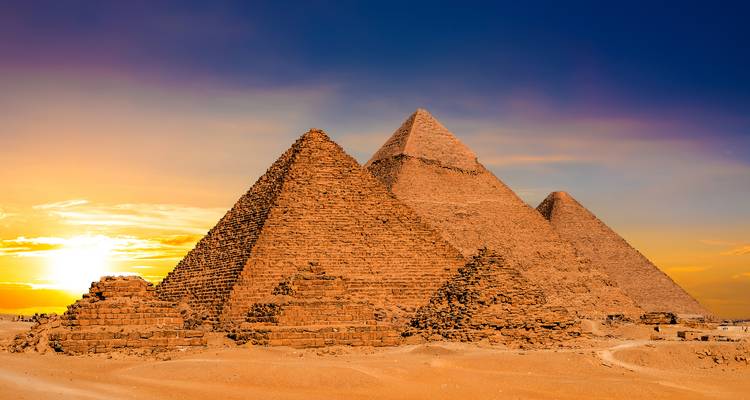 The three Great Pyramids of Giza rise from golden desert sands beneath a dramatic sunset sky.
