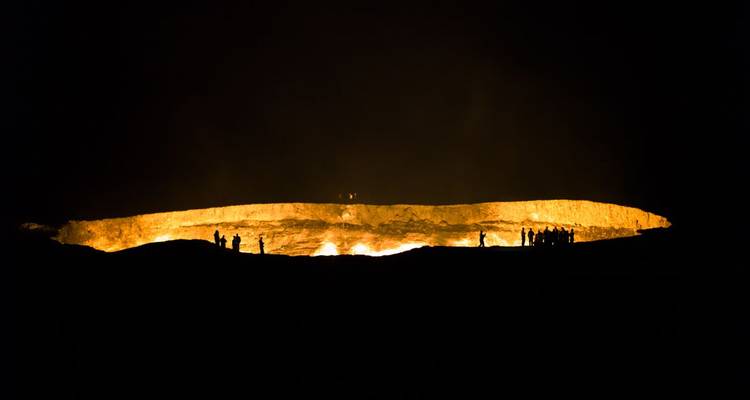 Large fire crater emitting flames at night with silhouettes of people.