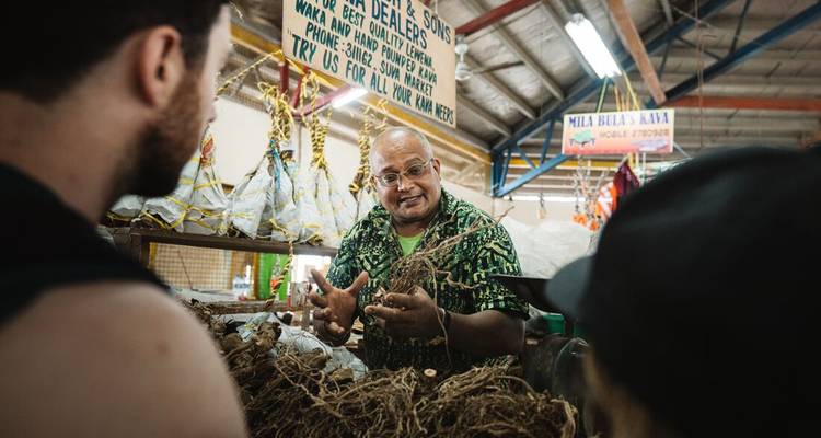 Un guide fidjien local explique avec enthousiasme des fagots de racines de kava aux voyageurs dans un marché animé.