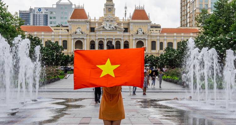 A person holding a Vietnam flag with a historical building and fountains.