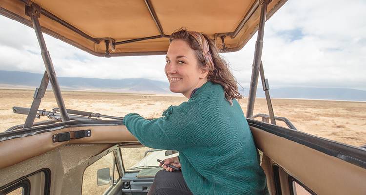 Viajero sonriente de pie asomado por el techo abierto de una jeepeta de safari con el paisaje de la sabana de fondo