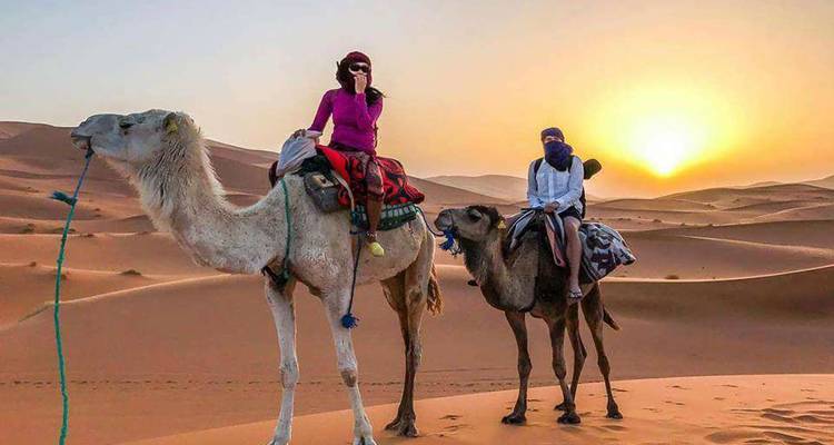 Two people riding camels in the desert at sunset.
