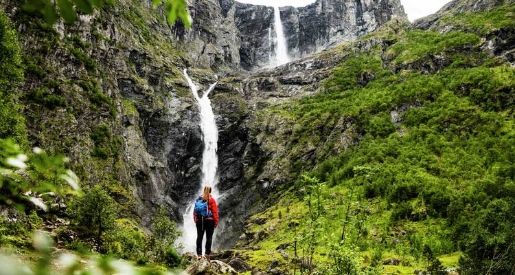 Person bewundert einen Wasserfall in einer üppigen Berglandschaft.