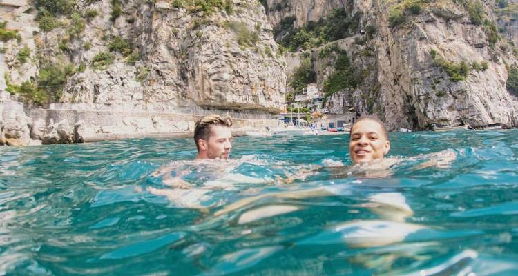 Two young men swim in clear blue coastal water beneath towering limestone cliffs.