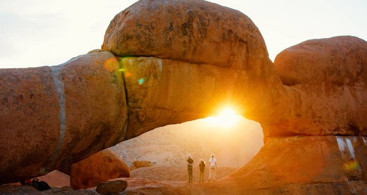 Vue du coucher de soleil avec des personnes debout sous une arche rocheuse naturelle.