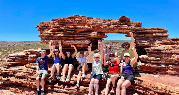 Grupo de turistas sonriendo posando con las manos alzadas bajo un arco natural de arenisca roja bajo el sol del outback.
