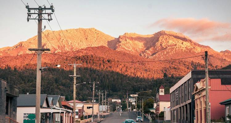 Rue d'une petite ville tasmanienne menant vers des montagnes accidentées et ensoleillées au crépuscule avec des lignes électriques au premier plan.