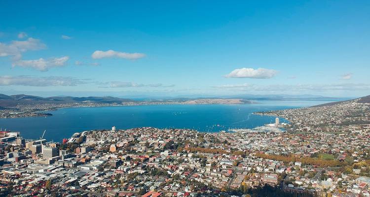 Vue aérienne panoramique de la ville de Hobart et de son port aux eaux bleu profond, encadrés par des collines lointaines sous un ciel dégagé.