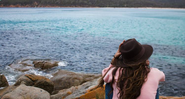 Jeune femme portant un chapeau assise sur des rochers côtiers contemplant la mer bleue étincelante et le littoral.